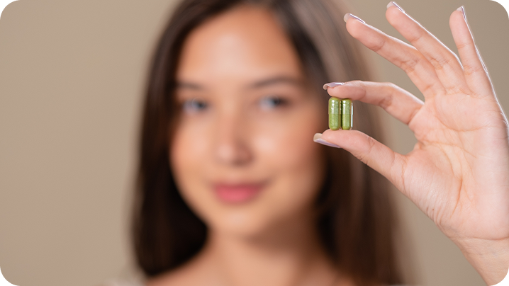 Mulher segura duas cápsulas verdes entre o polegar e o indicador, com a mão em primeiro plano e o rosto sorrindo suavemente ao fundo, desfocado.